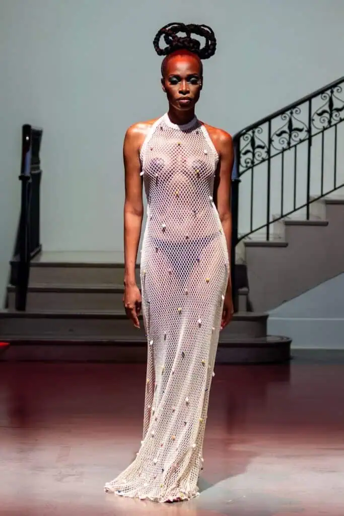 A model walks inside on a maroon floor, wearing a sheer, white, netted floor-length gown by emerging designers, adorned with small embellishments, with sculptural braided hair and dramatic makeup, standing in front of a staircase.