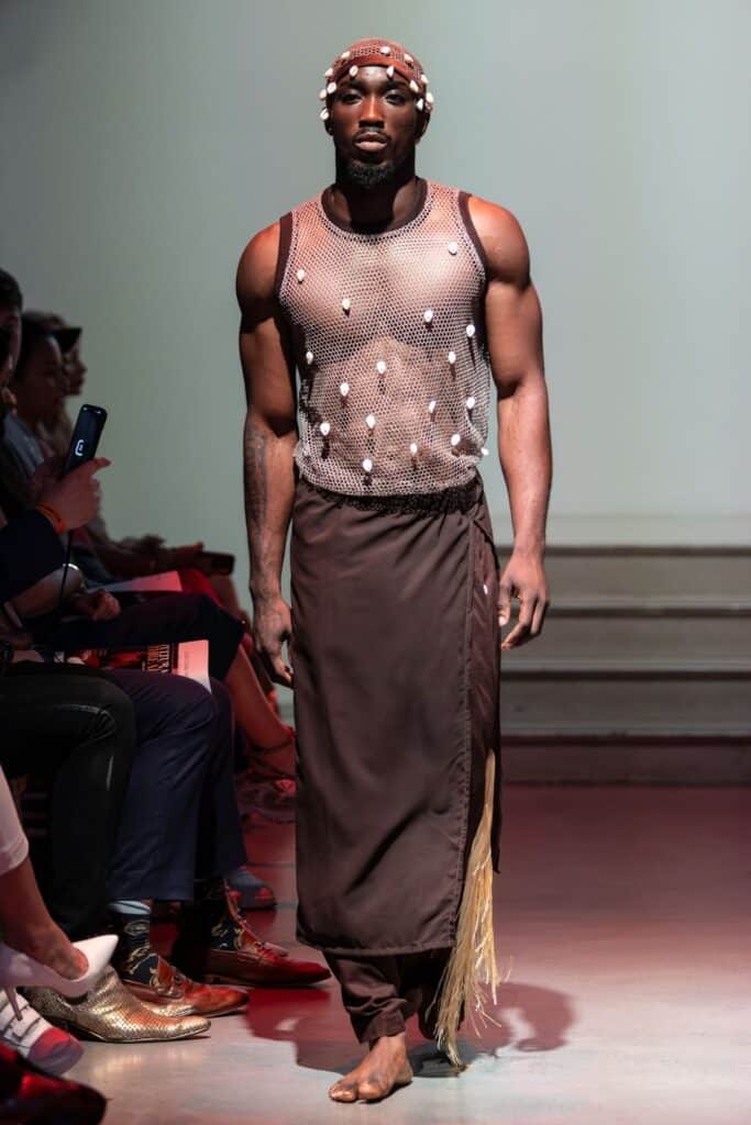 A model walks a runway for emerging designers, wearing a sleeveless mesh top adorned with shells, a long brown skirt with fringe details, and a beaded headpiece, barefoot, with an audience seated on either side.