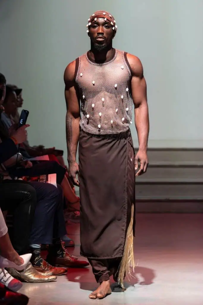 A model walks a runway for emerging designers, wearing a sleeveless mesh top adorned with shells, a long brown skirt with fringe details, and a beaded headpiece, barefoot, with an audience seated on either side.