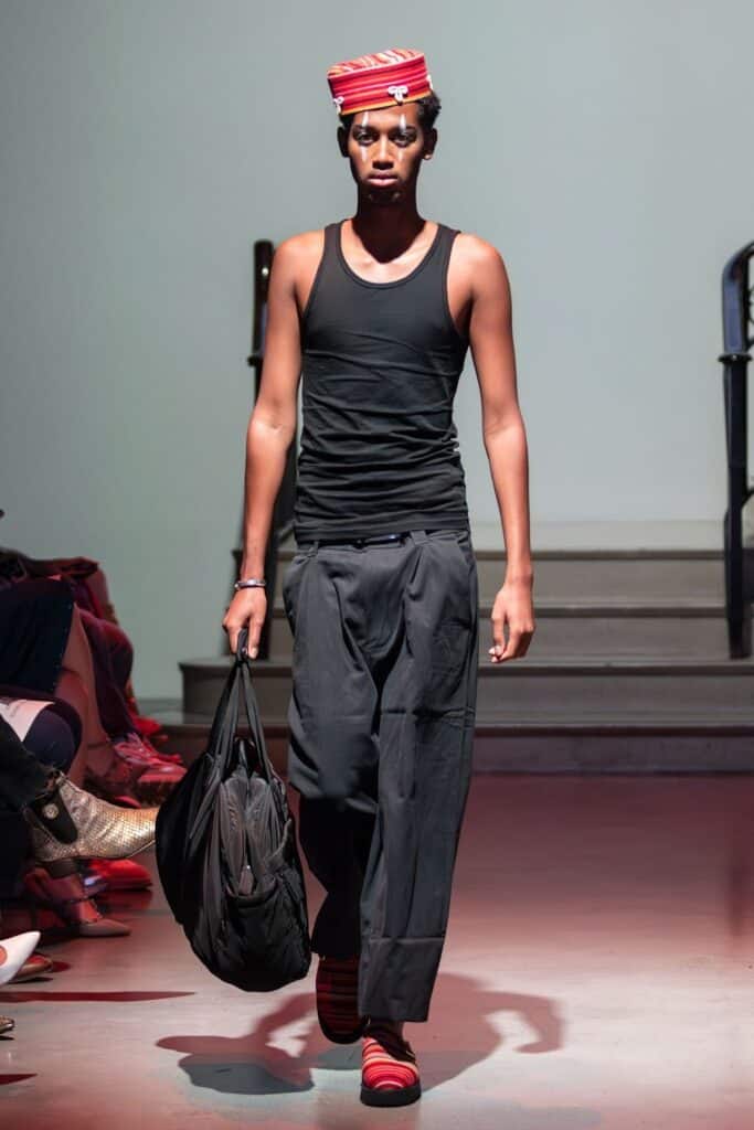 A model walks a runway for emerging designers, wearing a red striped hat, a black tank top, loose black pants, and red striped shoes, carrying a large black bag.