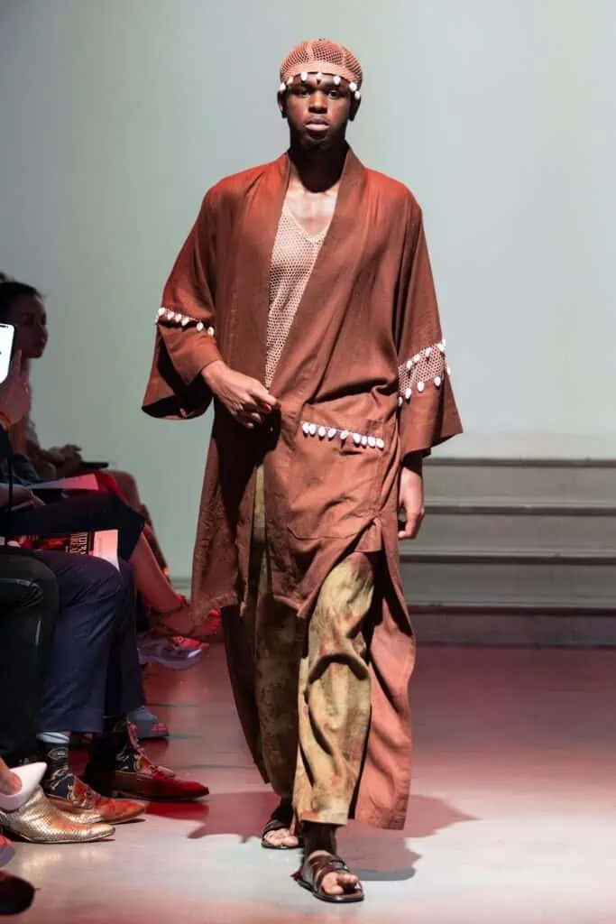 A model walks the runway in a brown, flowy robe with white detailing over a mesh shirt and patterned pants—an ensemble by emerging designers—paired with sandals and a beaded cap. The neutral background features soft lighting.