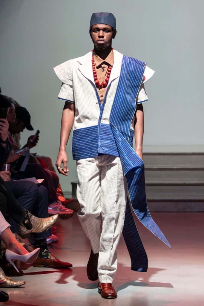 A male model walks the runway in a modern white and blue striped outfit by emerging designers, paired with a matching hat, brown undershirt, large red beaded necklace, and brown shoes. Spectators line the sides of the runway.