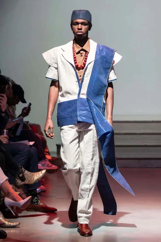 A male model walks the runway in a modern white and blue striped outfit by emerging designers, paired with a matching hat, brown undershirt, large red beaded necklace, and brown shoes. Spectators line the sides of the runway.