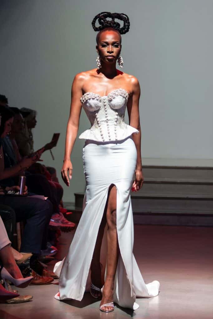 A model walks a runway in a white strapless gown with a sculpted, textured corset top and high slit skirt—an eye-catching look from emerging designers. She wears statement earrings and her hair is styled in intricate, twisted braids atop her head.