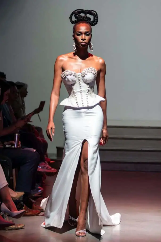 A model walks a runway in a white strapless gown with a sculpted, textured corset top and high slit skirt—an eye-catching look from emerging designers. She wears statement earrings and her hair is styled in intricate, twisted braids atop her head.