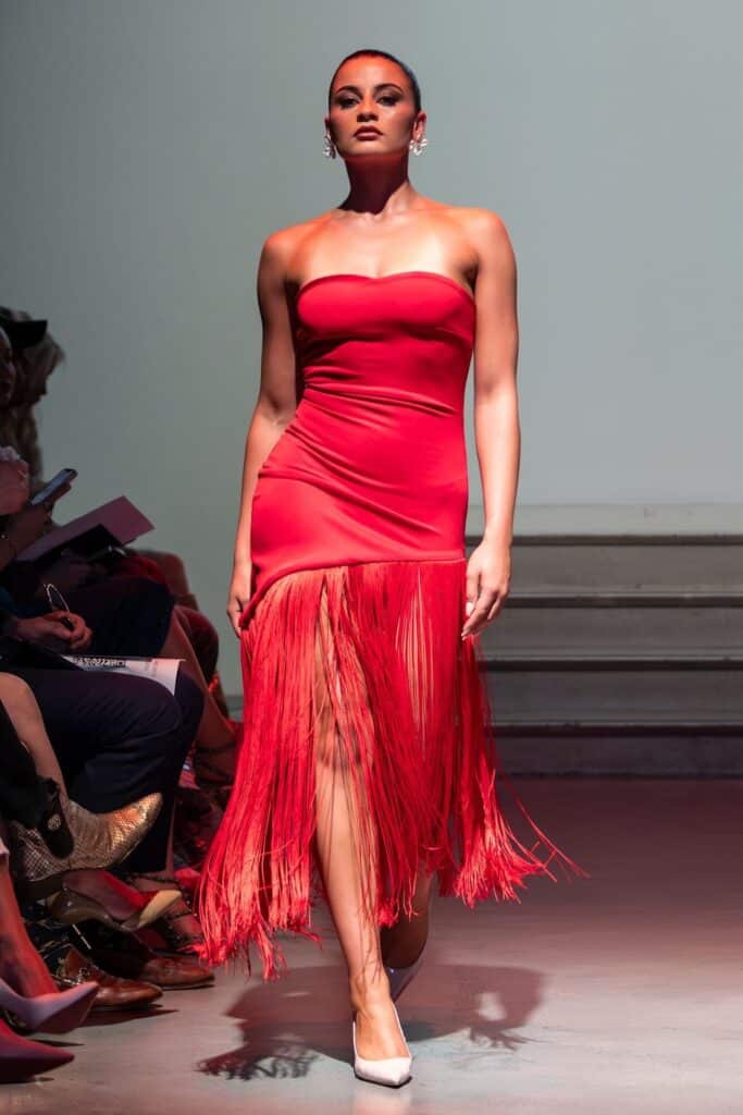 A model walks down a runway wearing a strapless, fitted red dress with a fringe skirt, white heels, and statement earrings—showcasing emerging designers. The background is plain with steps, and audience members are seated on either side.