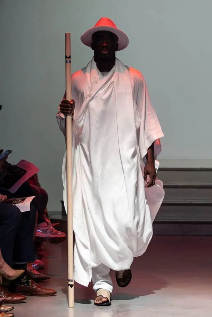 A model wearing a flowing white robe, wide-brimmed white hat, and sandals walks down the runway holding a tall staff. The minimal background and visible audience set the stage for emerging designers to showcase their creative vision.