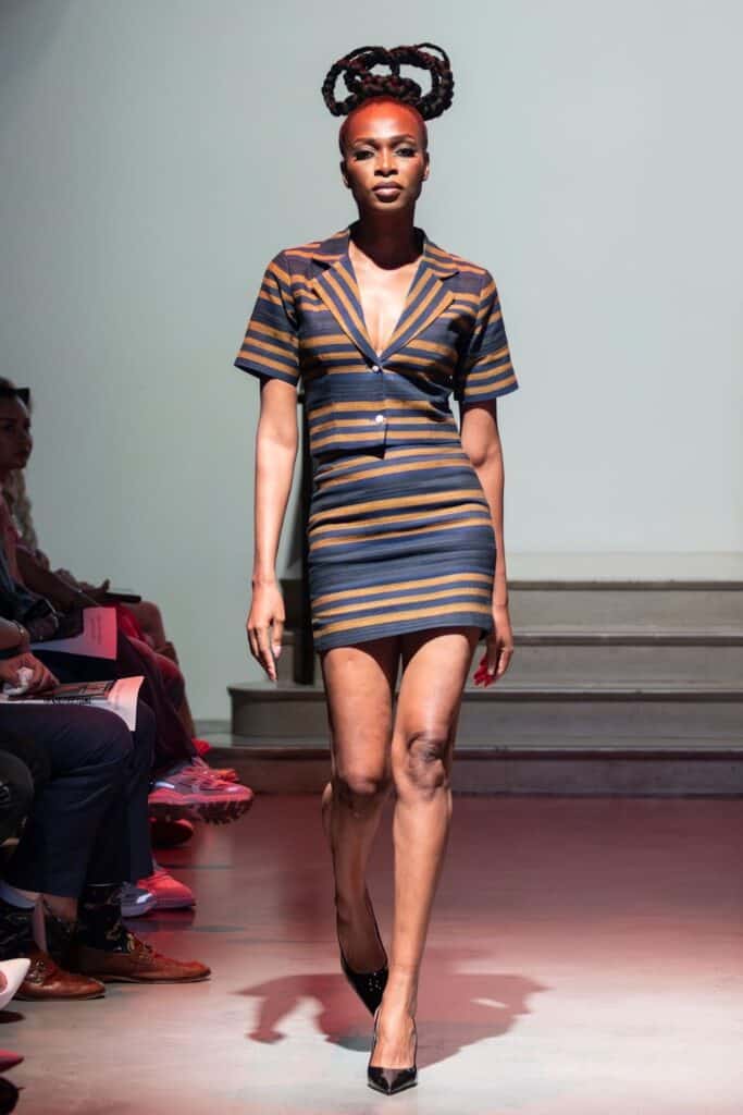 A model walks down a runway for emerging designers, wearing a short, fitted, striped dress with a deep V-neck and short sleeves. Her hair is styled in sculpted braids piled high, paired with chic black pointed heels.