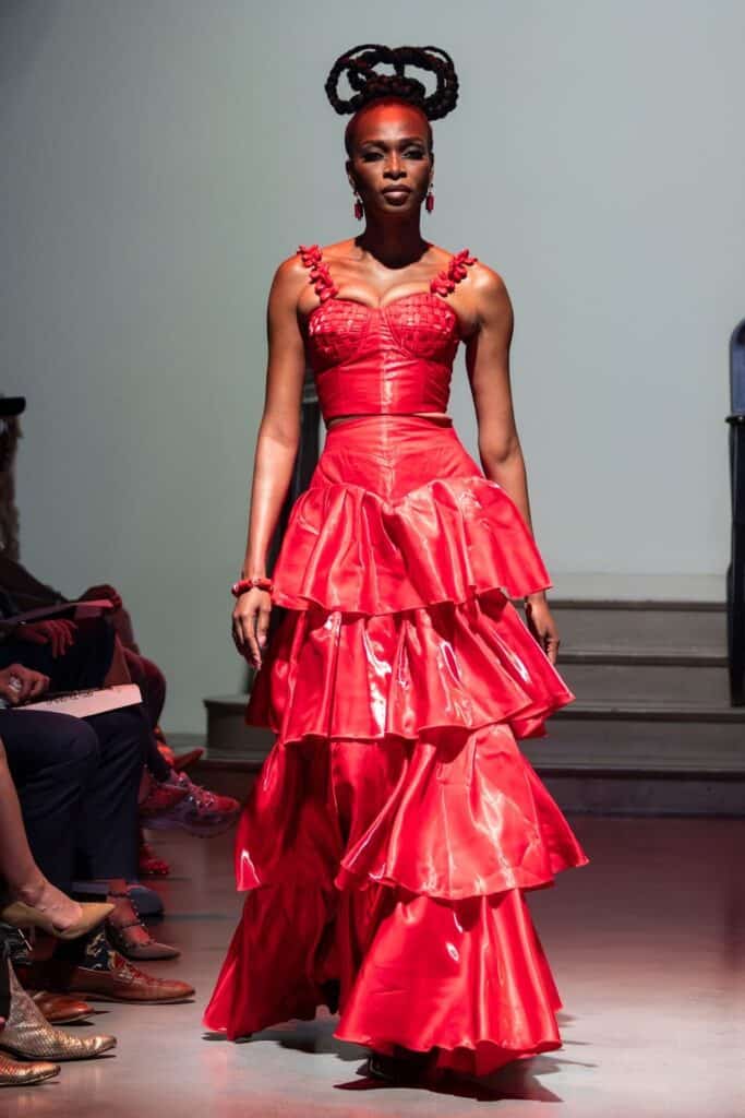 A model walks a runway for emerging designers in a bold, shiny red tiered ruffle gown with a fitted, corset-style bodice. Her hair is styled in an elaborate updo, and she accessorizes with red earrings and bracelets.