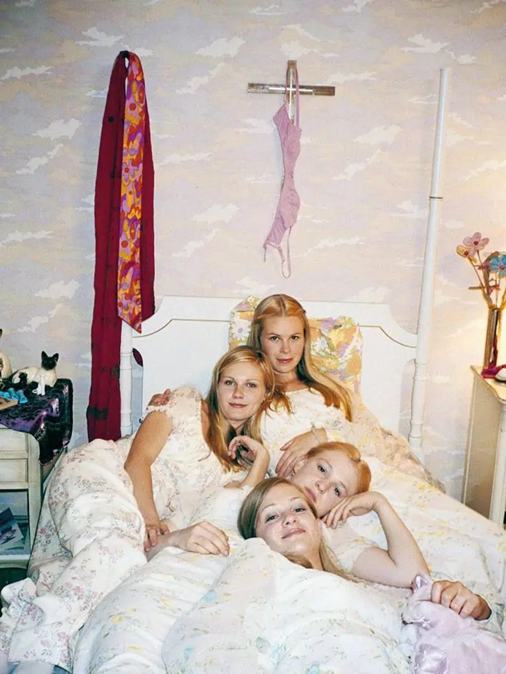 Archival photo from The Virgin Suicides showing four sisters lying together on a floral bed in a pastel bedroom, symbolizing innocence and melancholy.