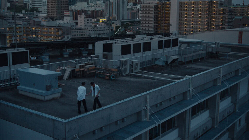 Two teenagers talk on a Tokyo rooftop at dusk in a still from Neo Sora’s film Happyend.