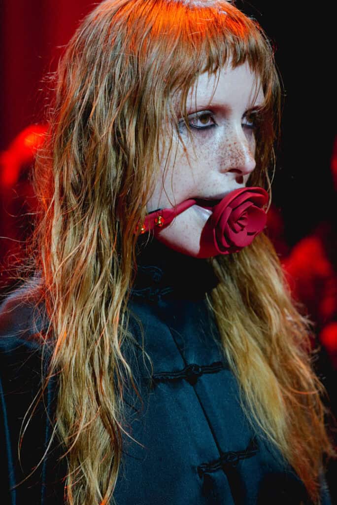 Close-up of a freckled model with wet hair in a black qipao-style dress, wearing a red rose bit-gag under red stage lights at Jenn Lee SS26.