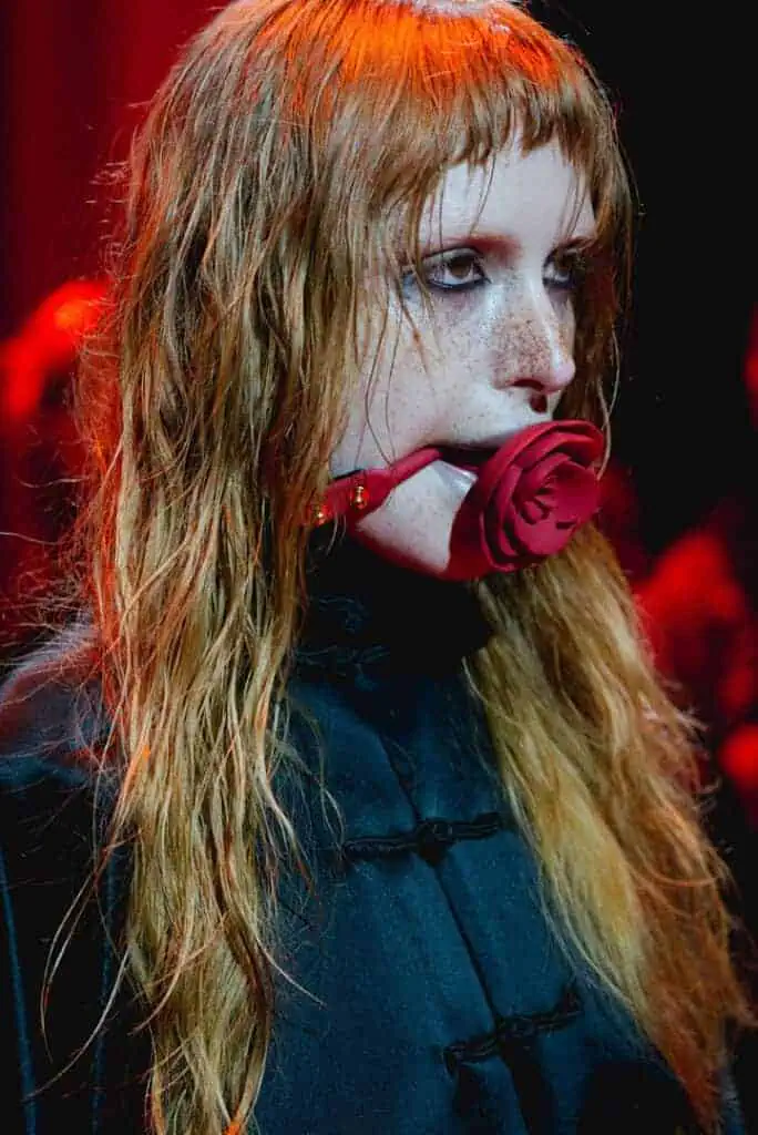 Close-up of a freckled model with wet hair in a black qipao-style dress, wearing a red rose bit-gag under red stage lights at Jenn Lee SS26.