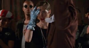 Party Girl (1995) scene where the protagonist wears a single glove while addressing another character, framed by nightlife associates in dim dramatic lighting.