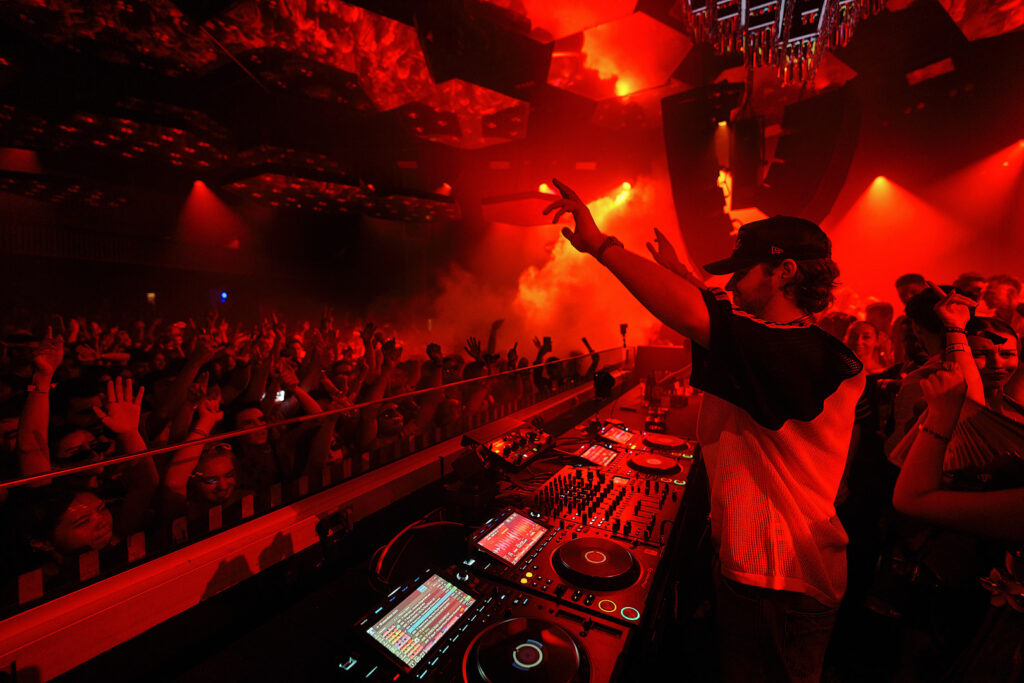 Mau P raising his hand behind the DJ booth as the crowd dances under red lights at Pacha Ibiza, photograph courtesy of the artist for Cold Magazine’s feature on “Tesla”.