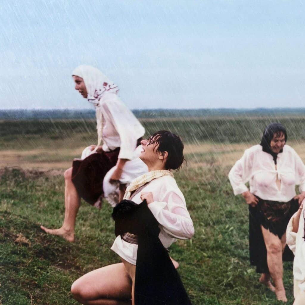 Performers in Marina Abramović’s Balkan Erotic Epic moving through a grassy field in ritual gestures, wearing traditional-style blouses and skirts in the rain.