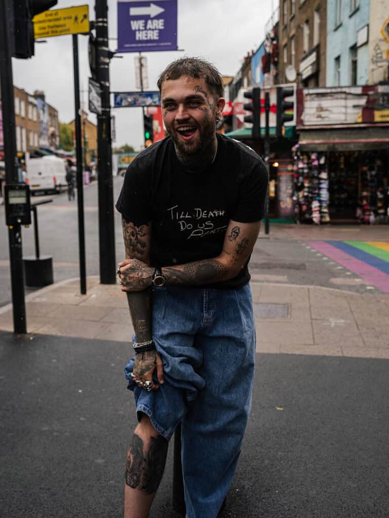 Portrait of Mason Newman smiling on a Camden street, showing his tattooed arms and wearing a black ‘Till Death Do Us Part’ T-shirt.