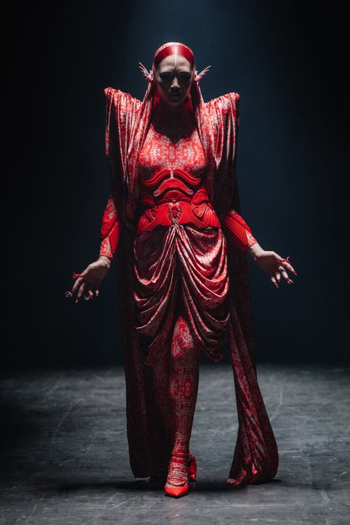 Model wearing a culptural red garment from the Hengdi Wang x STIM Precious Metals collaboration, featuring layered organic drapery and armor-like construction.