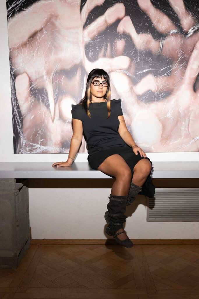Cate Colapietro seated in front of a large blurred photographic print featuring intertwined bodies at Cate Colapietro’s “Breath Not Found” exhibition in Milan.