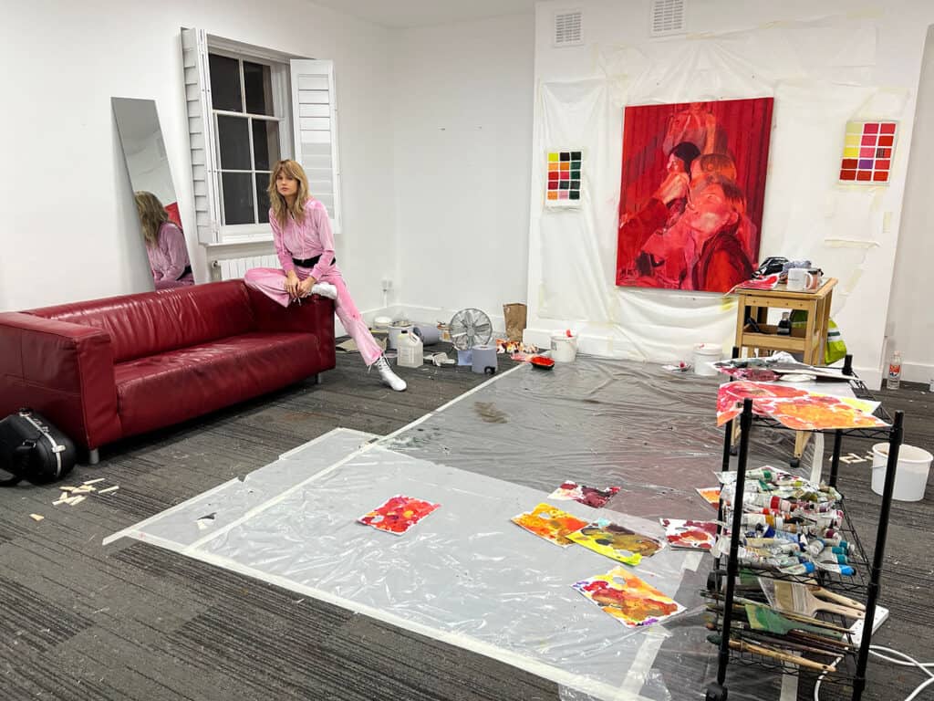 Studio view of Nana Wolke’s creative process for “Nothing Left to Want,” with Nana seated on a red sofa and multiple works in progress laid out across the floor.