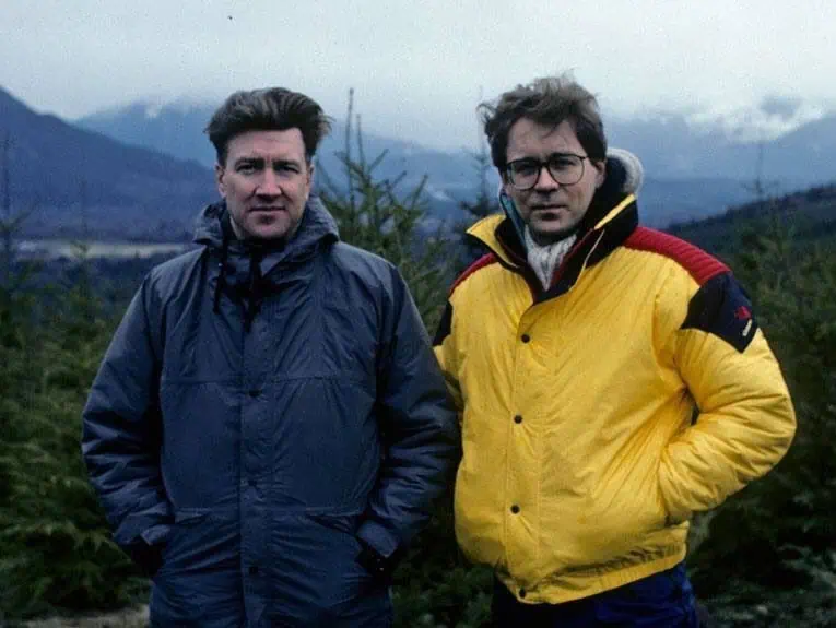 Mark Frost and David Lynch on the set of Twin Peaks, wearing winter jackets in the misty Pacific Northwest forest, reflecting the show’s mysterious and surreal atmosphere.