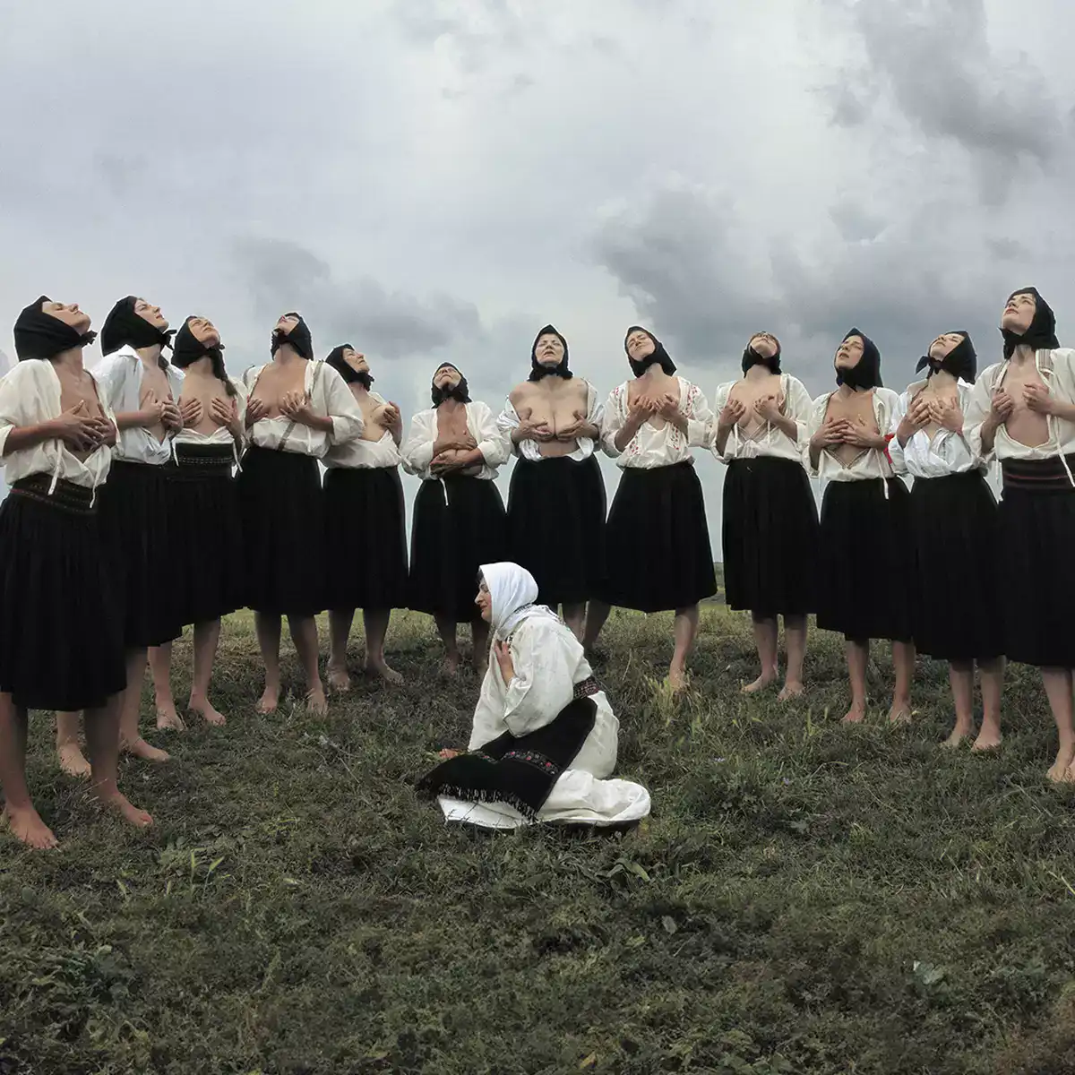 Outdoor ritual sequence from Balkan Erotic Epic, where a circle of performers stand in unison while one figure kneels in the grass.