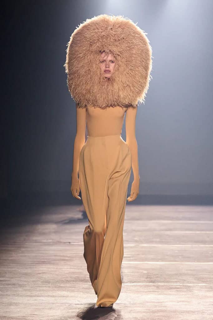 Mugler SS26 full-length beige bodysuit styled with oversized sculptural fur collar.
