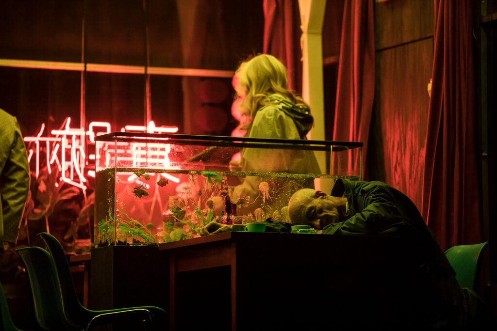 Performer gazing at a glass display filled with figurines under pink neon light, scene from Łukasz Twarkowski’s “ROHTKO”, photographed by Artūrs Pavlovs for Cold Magazine.