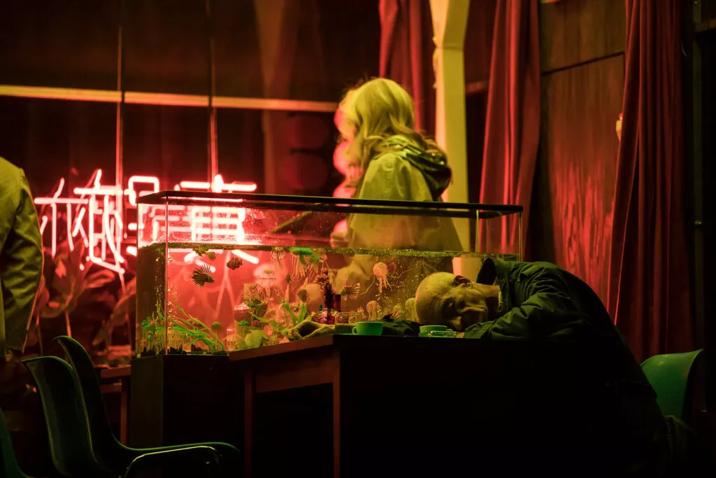 Performer gazing at a glass display filled with figurines under pink neon light, scene from Łukasz Twarkowski’s “ROHTKO”, photographed by Artūrs Pavlovs for Cold Magazine.