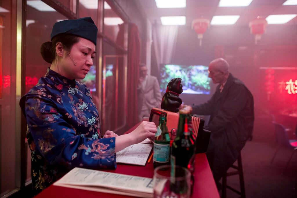 Scene from Łukasz Twarkowski’s “ROHTKO” showing a performer in traditional Chinese attire interacting with a customer at a counter, captured by Artūrs Pavlovs for Cold Magazine.
