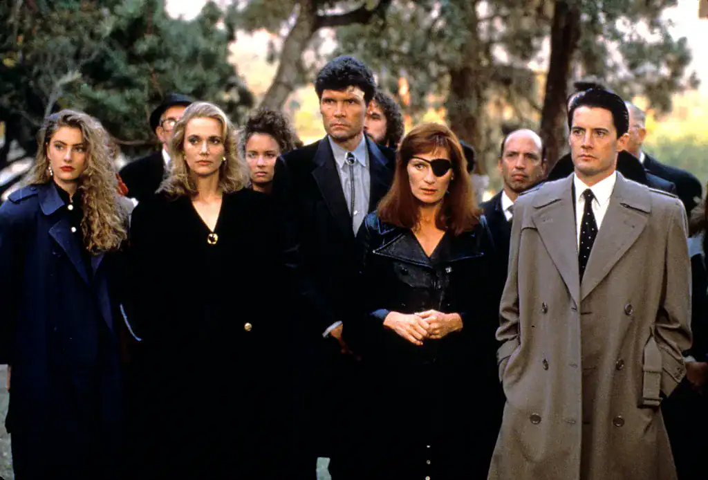 Group shot of characters from Twin Peaks standing together in outdoor daylight, dressed in dark coats.