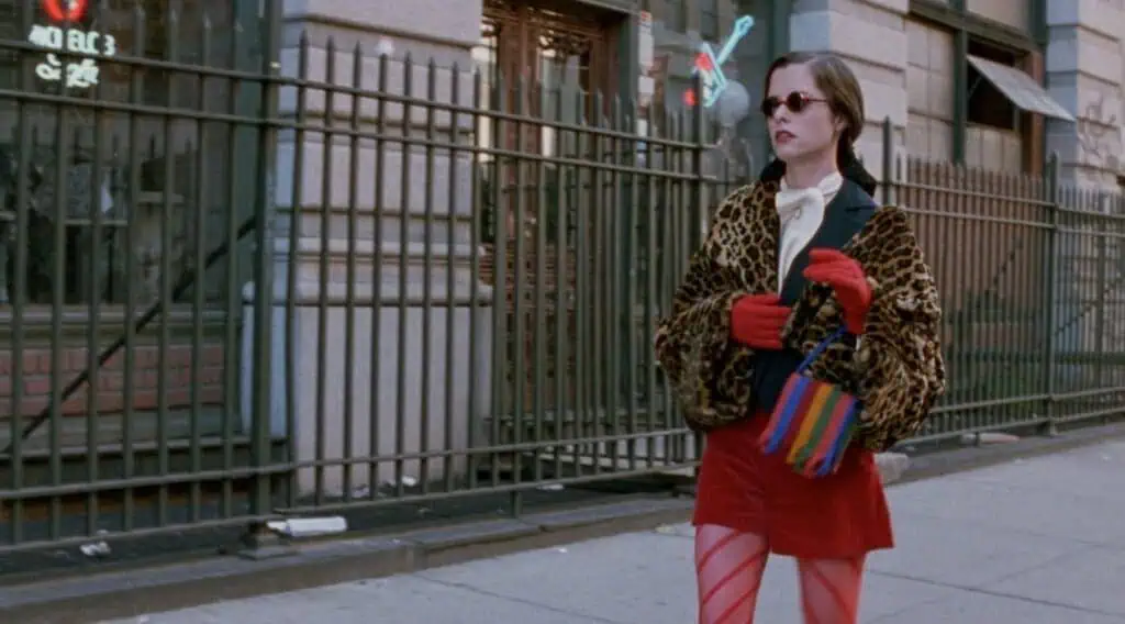 A Parker Posey in "Party Girl" wearing a leopard print jacket, red gloves, red skirt, and striped tights struts down a city sidewalk with a colorful bag. A metal fence and building with windows set the backdrop.