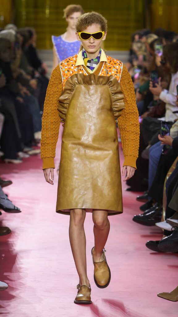 Miu Miu SS26 runway look with honey leather apron and textured orange sweater.