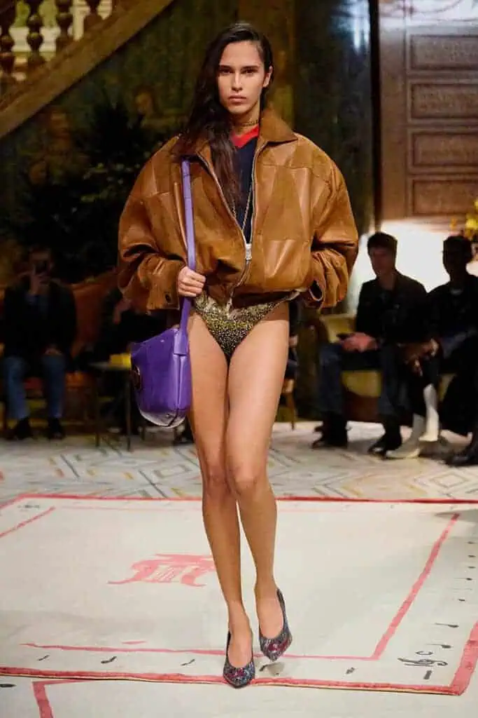 Model in oversized brown leather bomber jacket, metallic briefs, and purple shoulder bag from Versace Spring/Summer 2026 collection.