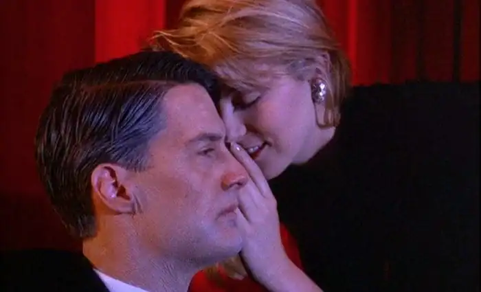 Scene from Twin Peaks where one character leans in to whisper into another’s ear, framed in warm red lighting.