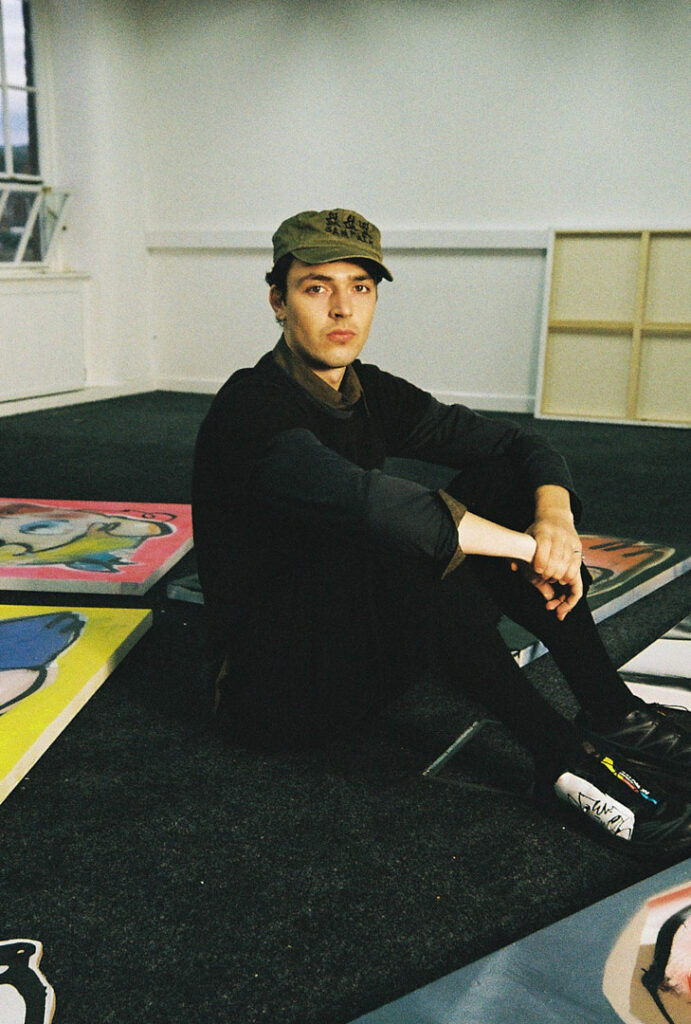 Artist Tomas Gittins seated on the studio floor beside his colourful, expressive character paintings.