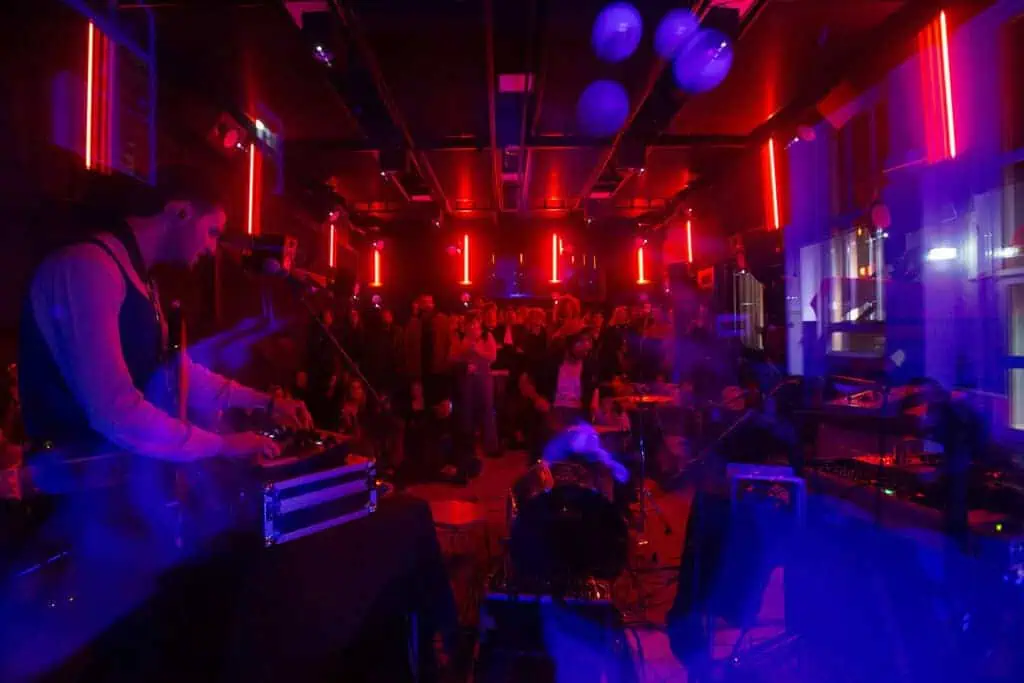 A musician playing synthesizers in a dark room lit by red and blue neon lights, with the audience gathered closely during a Linecheck performance.