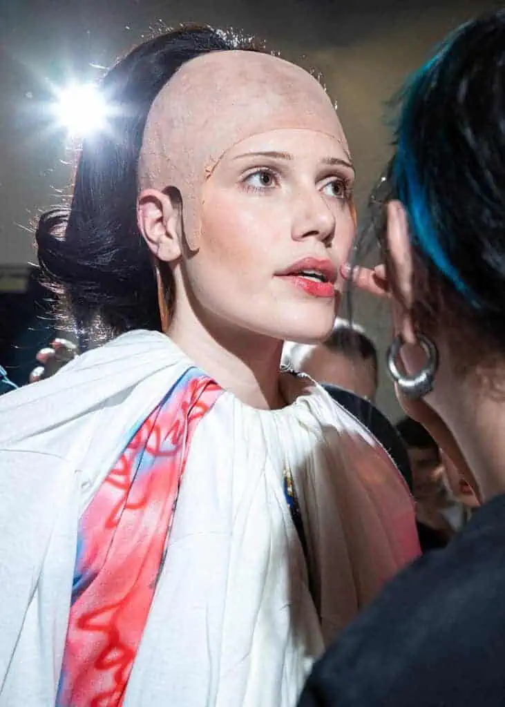 Backstage close-up of a model having makeup applied, wearing Matil Vanlint’s sculptural costume and prosthetic wig