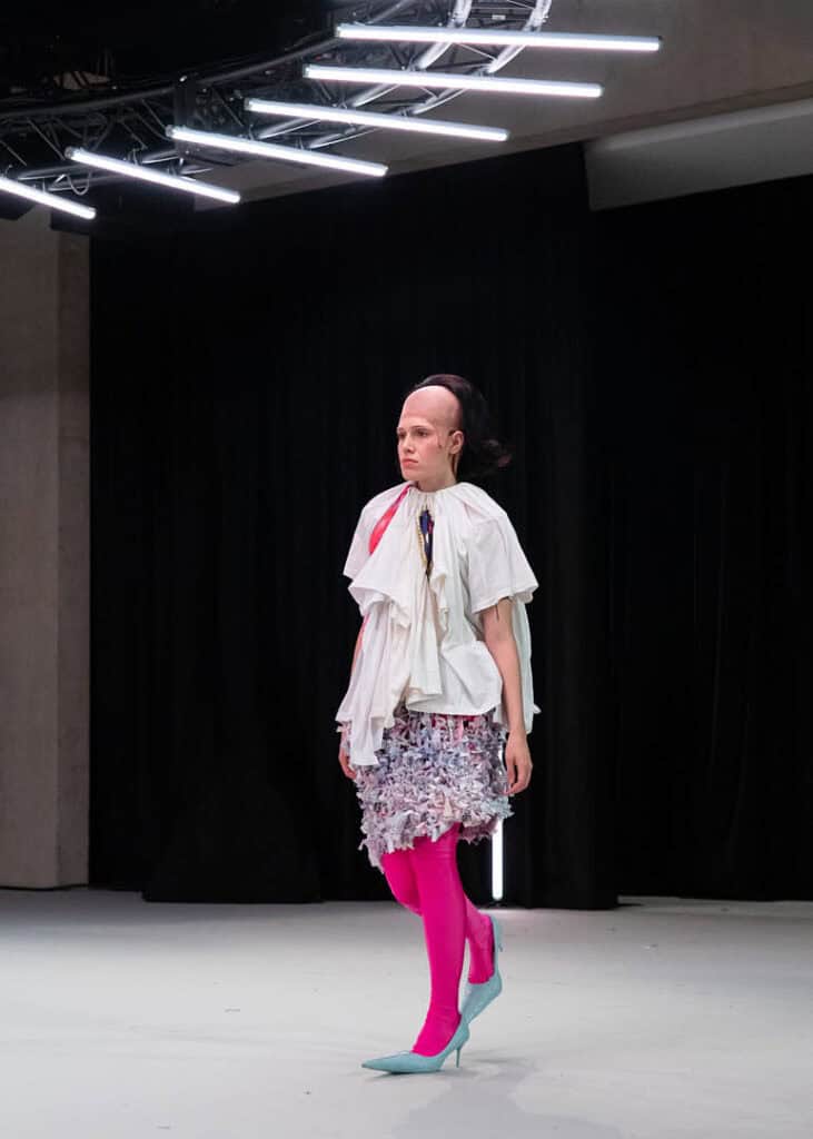 Model in pink tights and a deconstructed white top from Matil Vanlint’s V collection walking the runway.