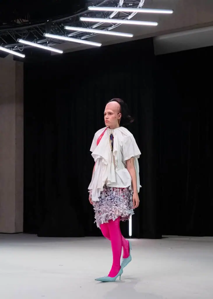 Model in pink tights and a deconstructed white top from Matil Vanlint’s V collection walking the runway.