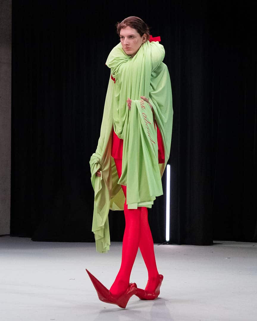 A model wearing an avant-garde creature-like outfit by Matil Vanlint, featuring flowing green fabric, red tights, and sculptural red shoes on the runway at HEAD Genève.