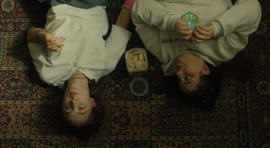 Two teenage girls lying on a patterned rug eating communion wafers in a scene from “Lady Bird” (2017).