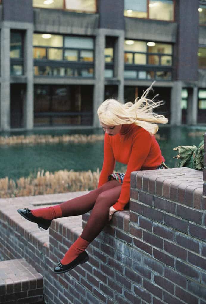 Dakota Warren sitting on a brick wall by the water, wearing a red outfit posing for The cold magazine.