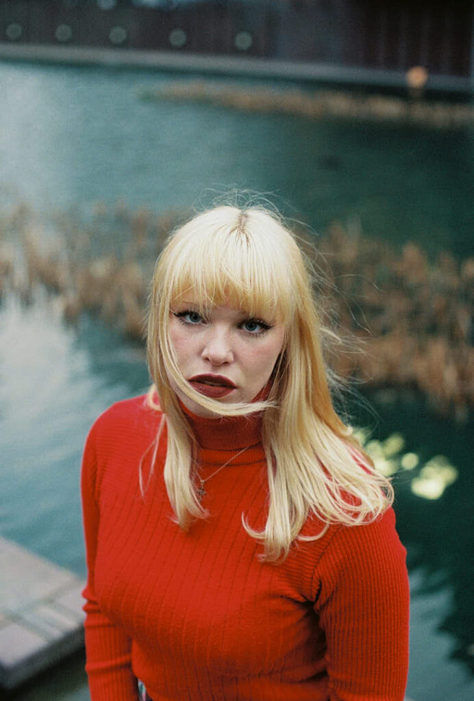 Portrait of Dakota Warren in a red turtleneck near a lakeside setting posing for The cold magazine.