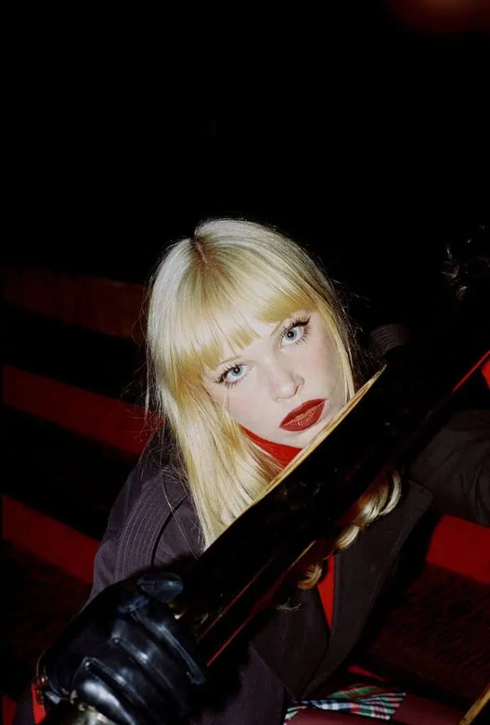 Flash-lit close-up of Dakota Warren with red lips holding a metallic railing posing for The cold magazine.