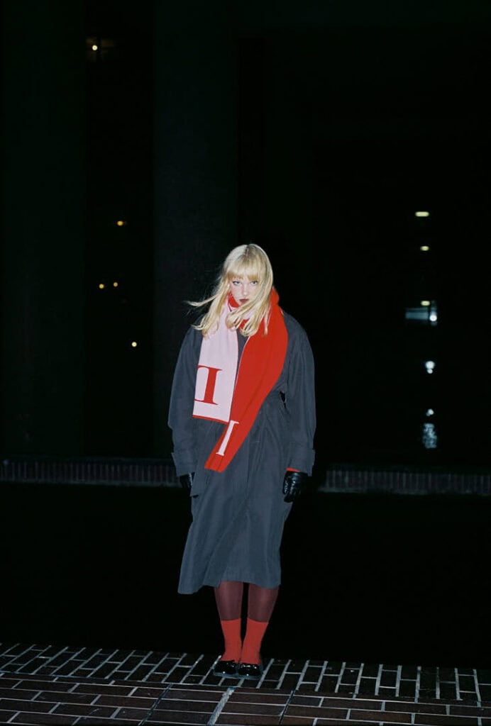 Dakota Warren in a dark urban setting wearing a long coat and red scarf at night posing for The cold magazine.