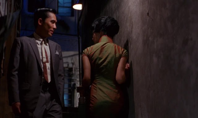 Man and woman passing each other in a narrow alley at night, with the woman carrying a thermos of noodles in a scene from “In the Mood for Love” (2000).