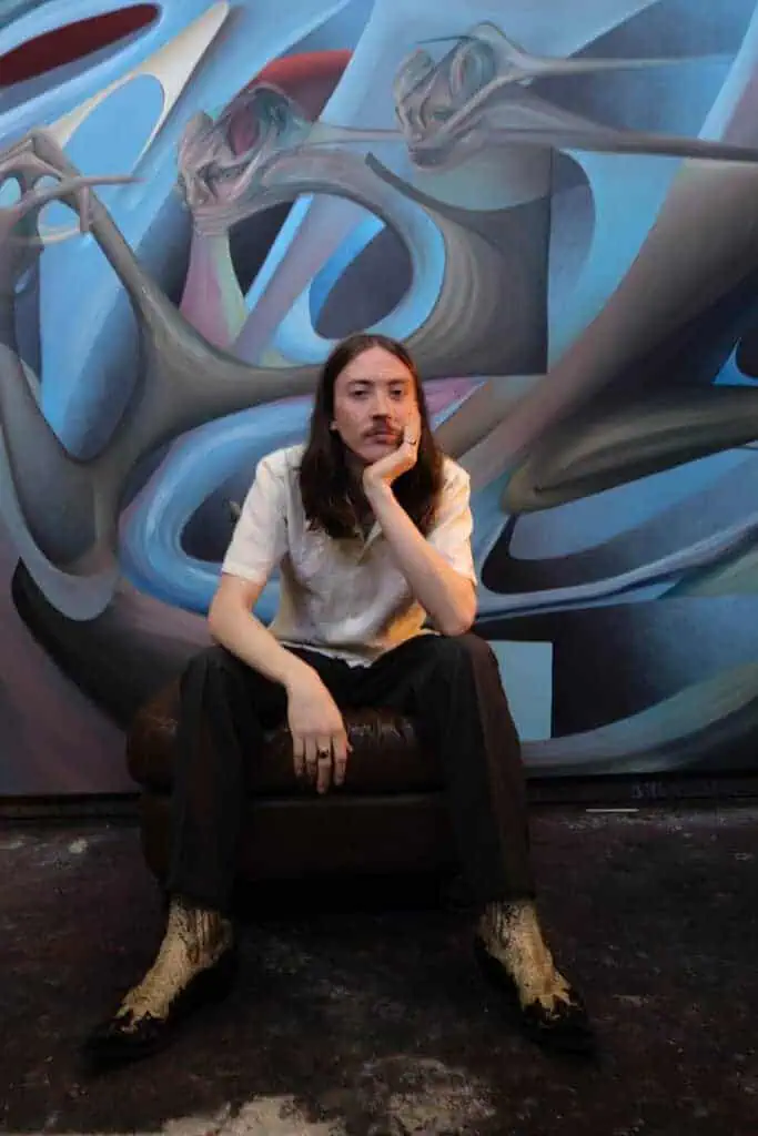 Portrait of Alfie Rouy in front of a surreal abstract painting with fluid, distorted forms and cool-toned hues, suggesting movement and introspection.