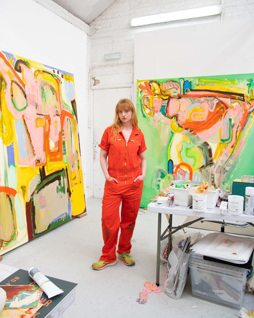 Portrait of artist Bunny Hennessey in a bright studio, standing between two large abstract canvases in vibrant colours, surrounded by paints and tools.