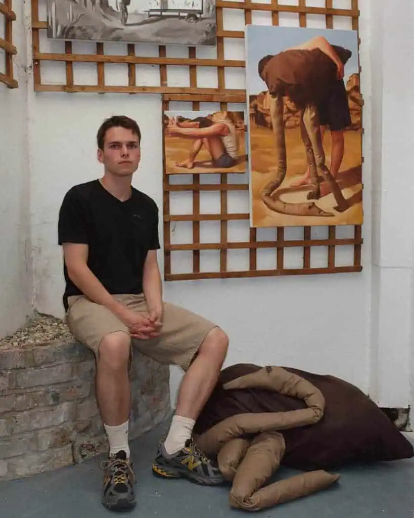 Photograph of artist Conor Quinn seated in his studio surrounded by figurative paintings exploring surrealism and material form.